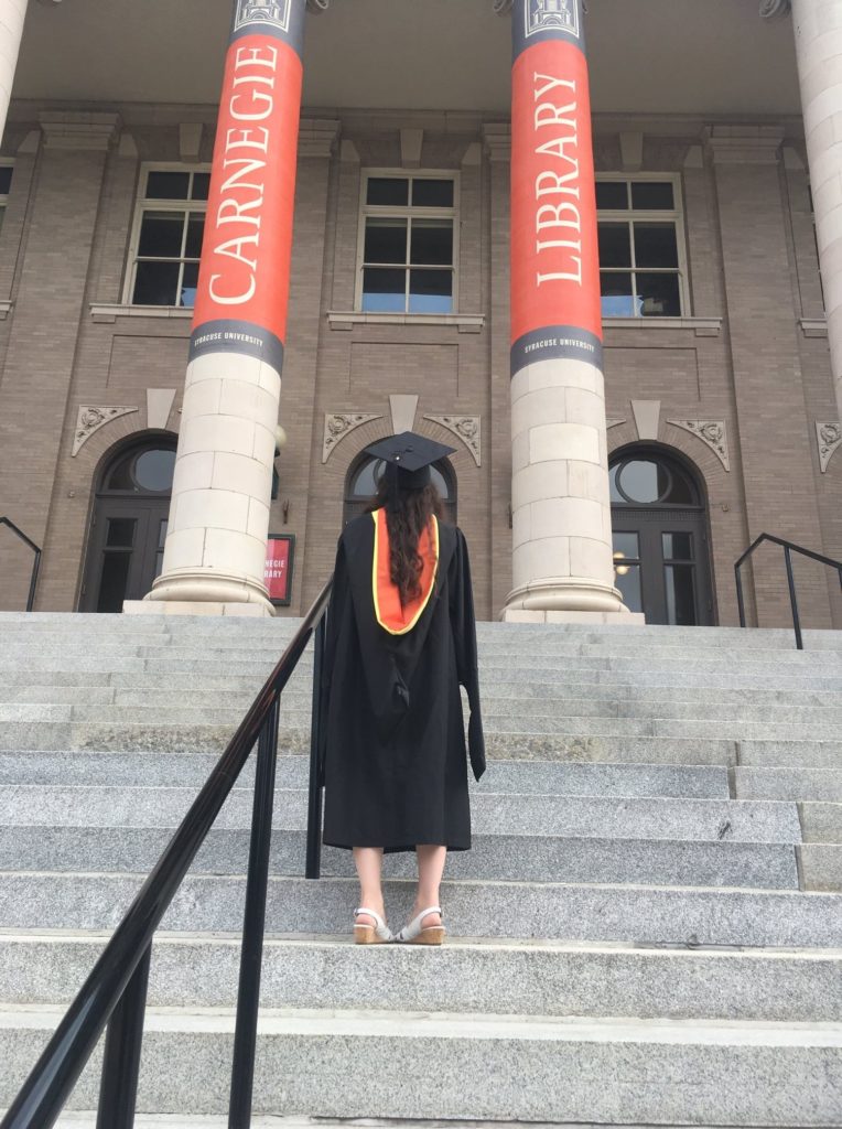 Vivian Teresa Tompkins in a graduation gown on the steps of a library, facing away from the camera.